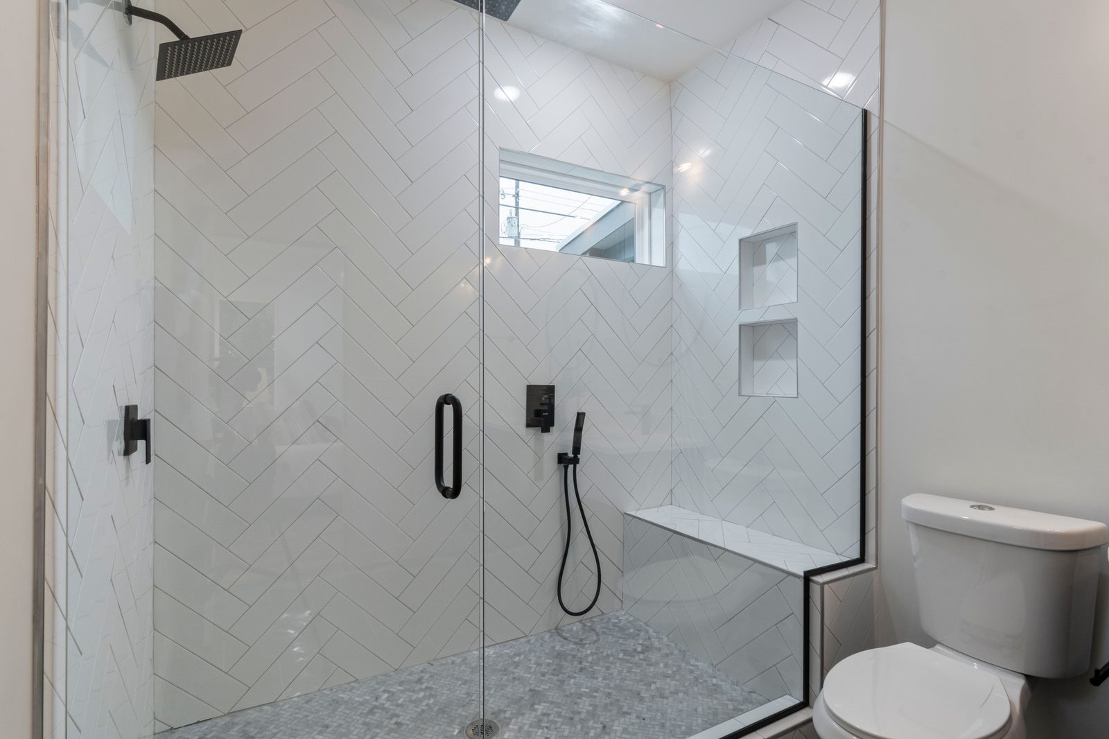 Frameless glass shower door on white herringbone tile with matte black hardware and bench, by Makibaki in Deerfield, IL